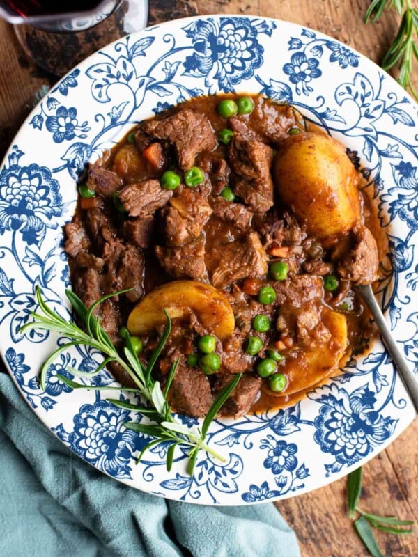 Italian beef stew with potatoes and peas in a blue and white floral bowl with rosemary and red wine at the side.
