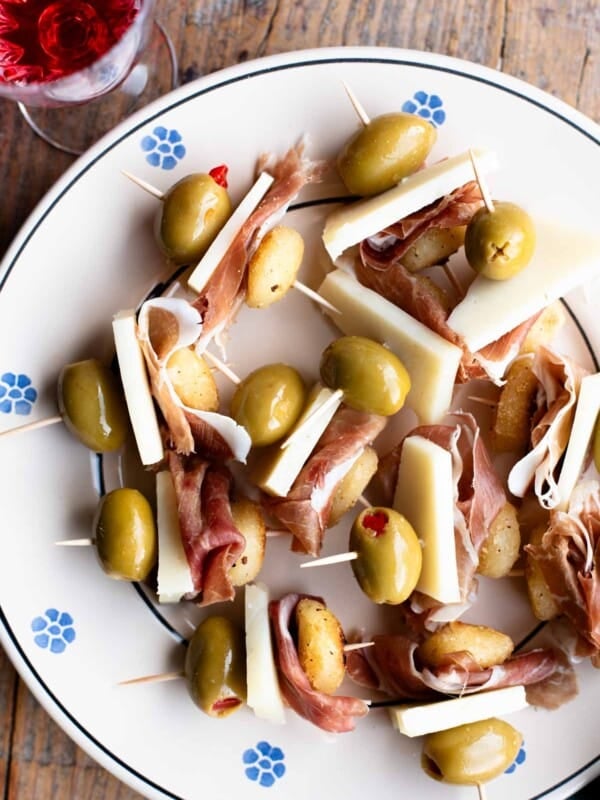 Little fried gnocchi appetizers with prosciutto, pecorino and olives in toothpicks sitting on a plate with a rustic wooden background.