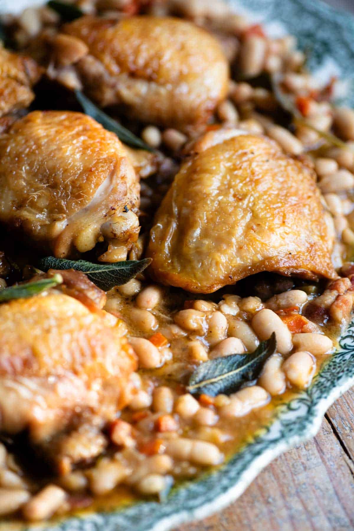 A close up of braised chicken on a serving plate with beans and sage.