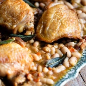 A close up of braised chicken thighs sitting on a plate with beans in sauce with sage and vegetables.