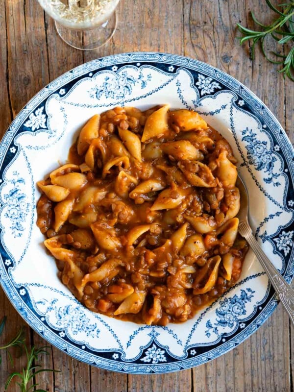 An overhead shot of pasta and lentils in a blue vintage style bowl on a rustic background with a glass of white wine at the side.