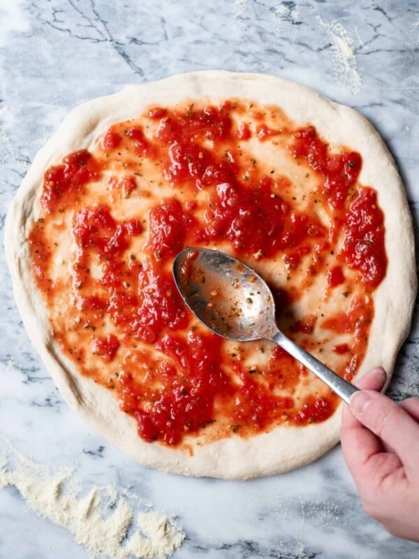 A pizza base on a marble work surface with tomato sauce getting spread on top.