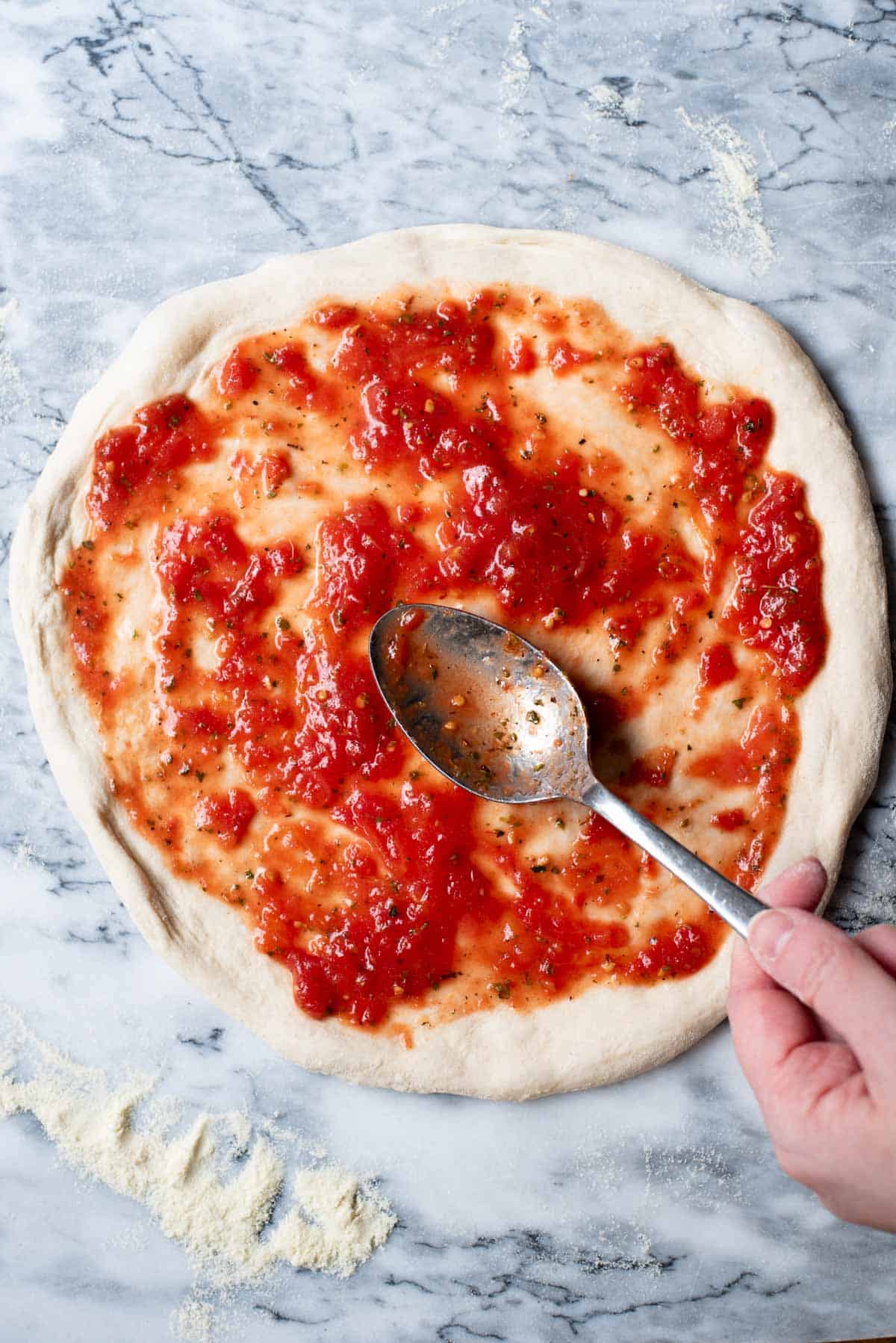 A pizza base on a marble work surface with tomato sauce getting spread on top.