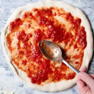 A close up of a pizza base getting tomato sauce spread on top before baking.