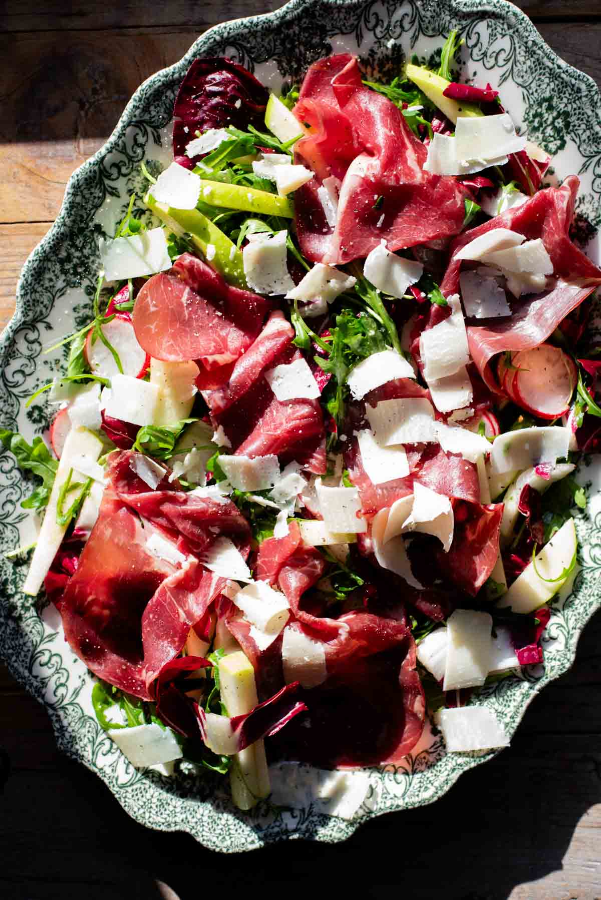 A salad with Bresaola, radicchio, radish and apple in direct sunlight on a large green and white platter.