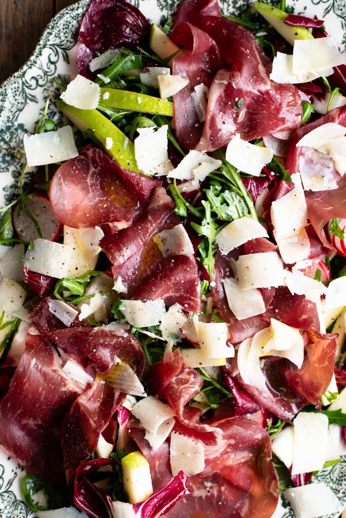 A close up of a salad on a green and white serving plate with bresaola, arugula, radicchio, radish and apple.