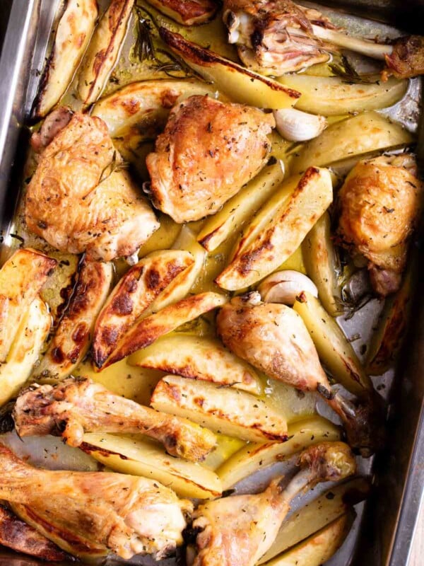 A close up overhead shot of chicken and potato wedges in a roasting tray with garlic and rosemary.