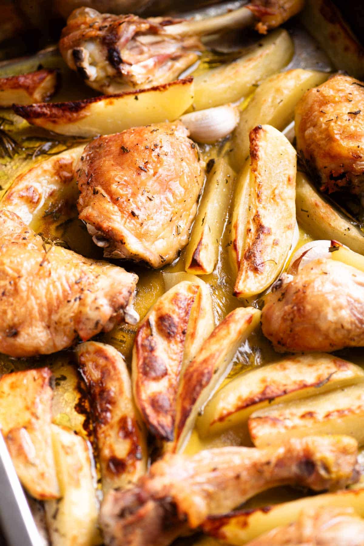 A close up side shot of roasted chicken thighs and drumsticks with potato wedges in a roasting tray.