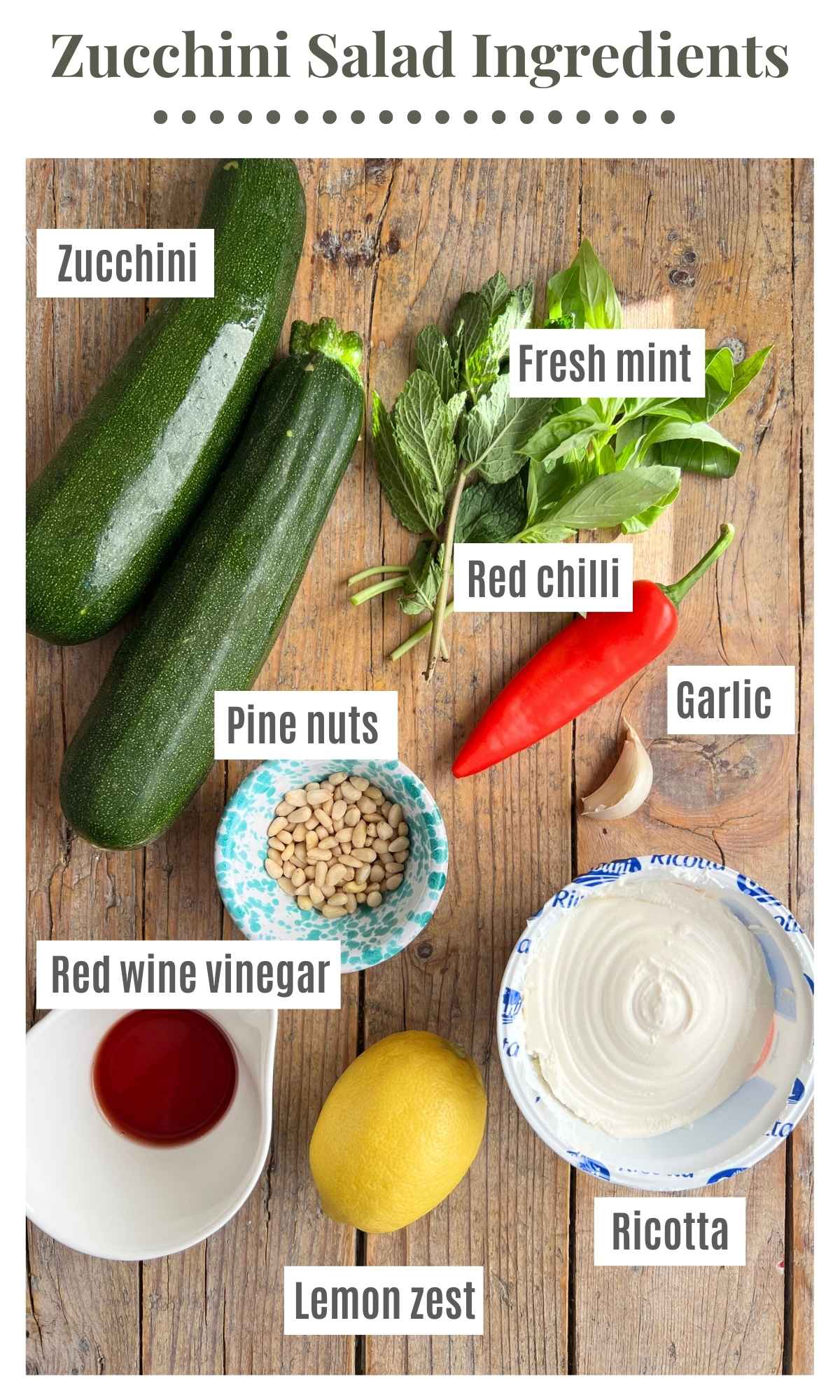 Zucchini, garlic, red chilli, pine nuts, red wine vinegar, ricotta, a whole lemon and mint laying on a rustic wooden surface.