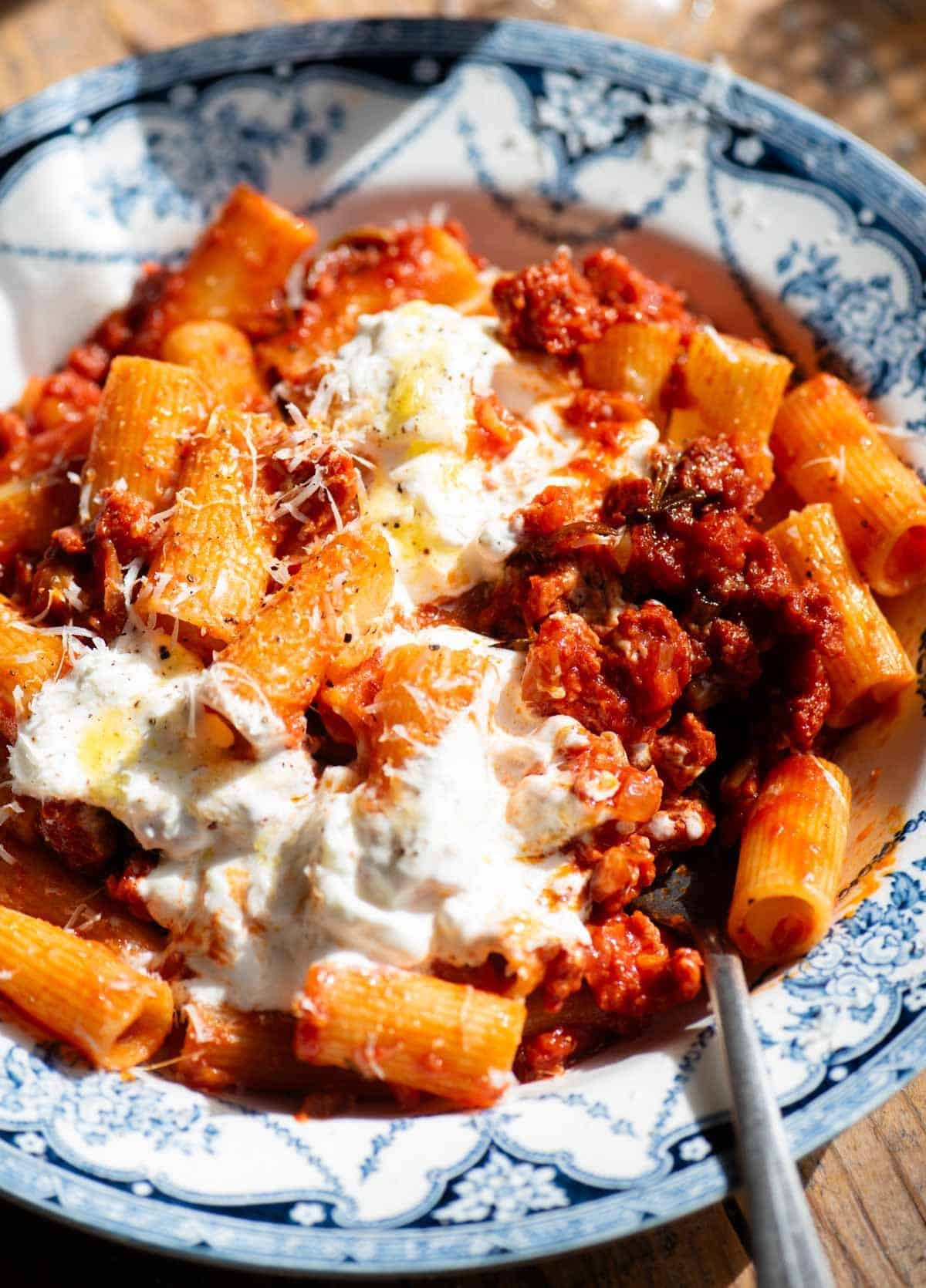 A close up of a spicy sausage ragu topped with stracciatella cheese in a blue and white bowl and a fork at the side.