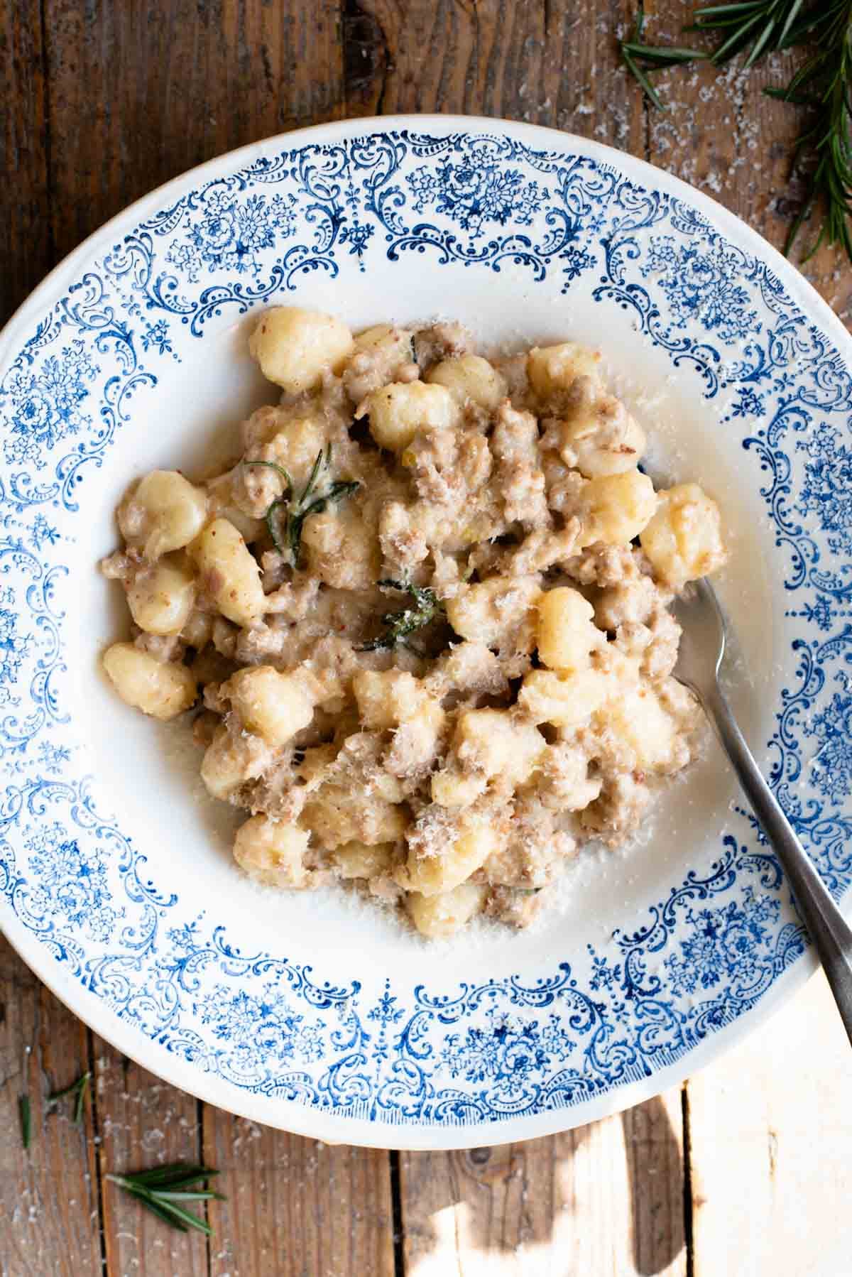 An overhead shot of gnocchi with a white creamy sausage ragu in a blue and white bowl on a rustic wooden surface.