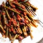 A close up of green beans with olives, capers and tomato on a white plate.