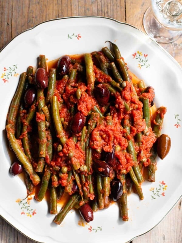 A close up of green beans in tomato sauce with olives and capers on a white floral plate and rustic wooden background.