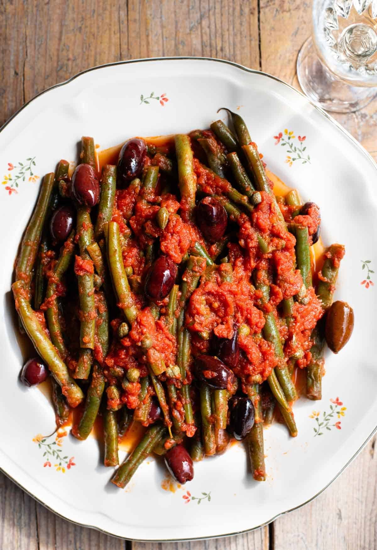 A close up of green beans in tomato sauce with olives and capers on a white floral plate and rustic wooden background.