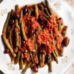 A close up of green beans in tomato sauce with olives and capers on a white plate.