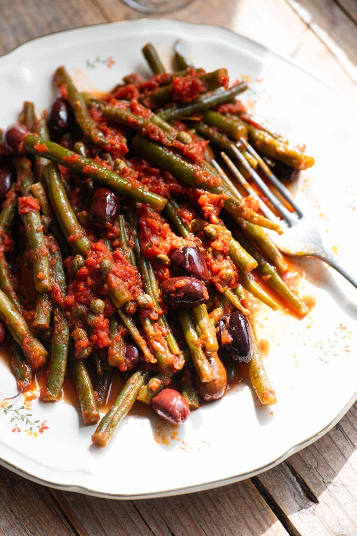 A side shot of green beans with tomatoes, olives and capers on a white plate with a fork at the side.