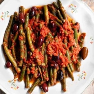 A close up of green beans in tomato sauce with olives and capers on a white plate.