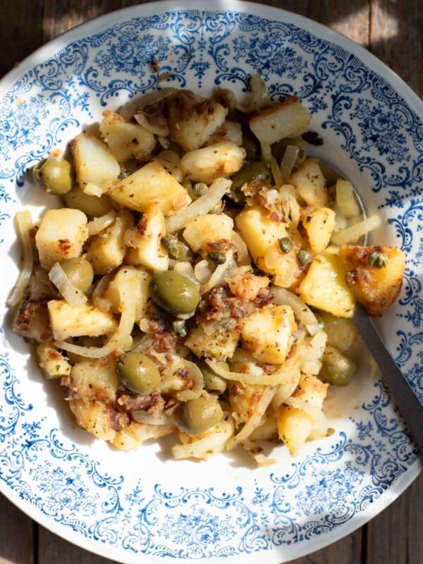 An overhead shot in direct sunlight of Sicilian potatoes in a blue bowl with olives and capers.