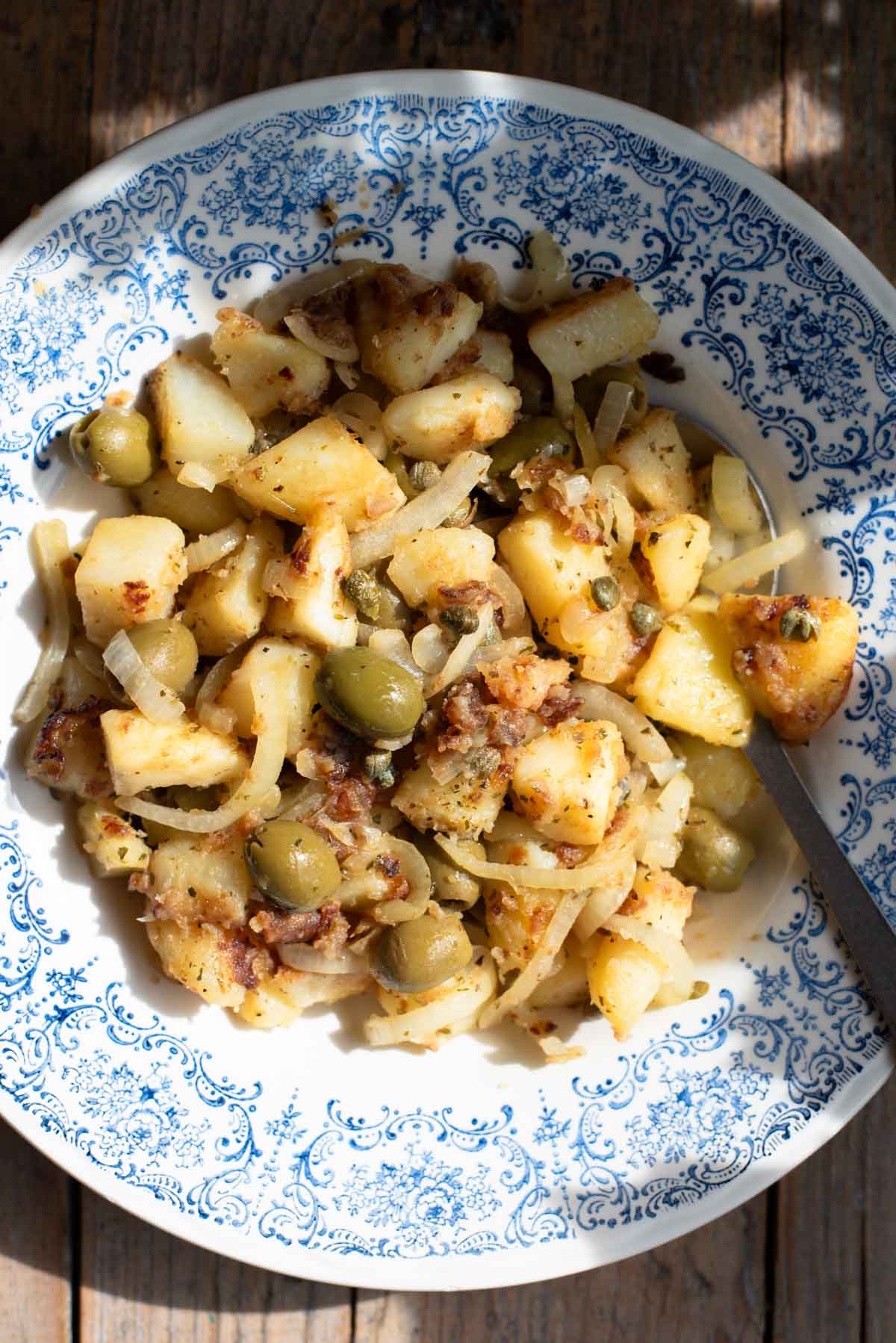 An overhead shot in direct sunlight of Sicilian potatoes in a blue bowl with olives and capers.
