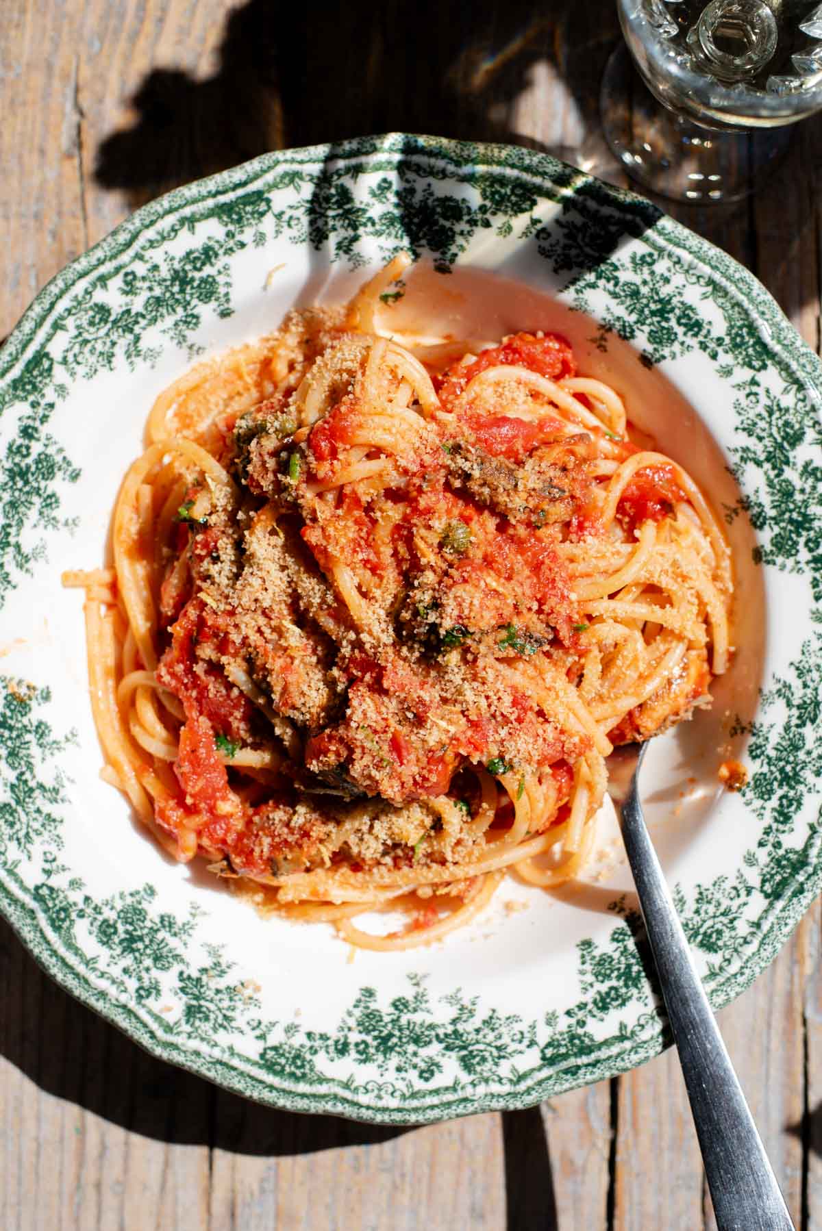 An overhead shot of spaghetti in a tomato sauce with sardines and breadcrumbs on top. The bowl is a vintage style green and white colour.
