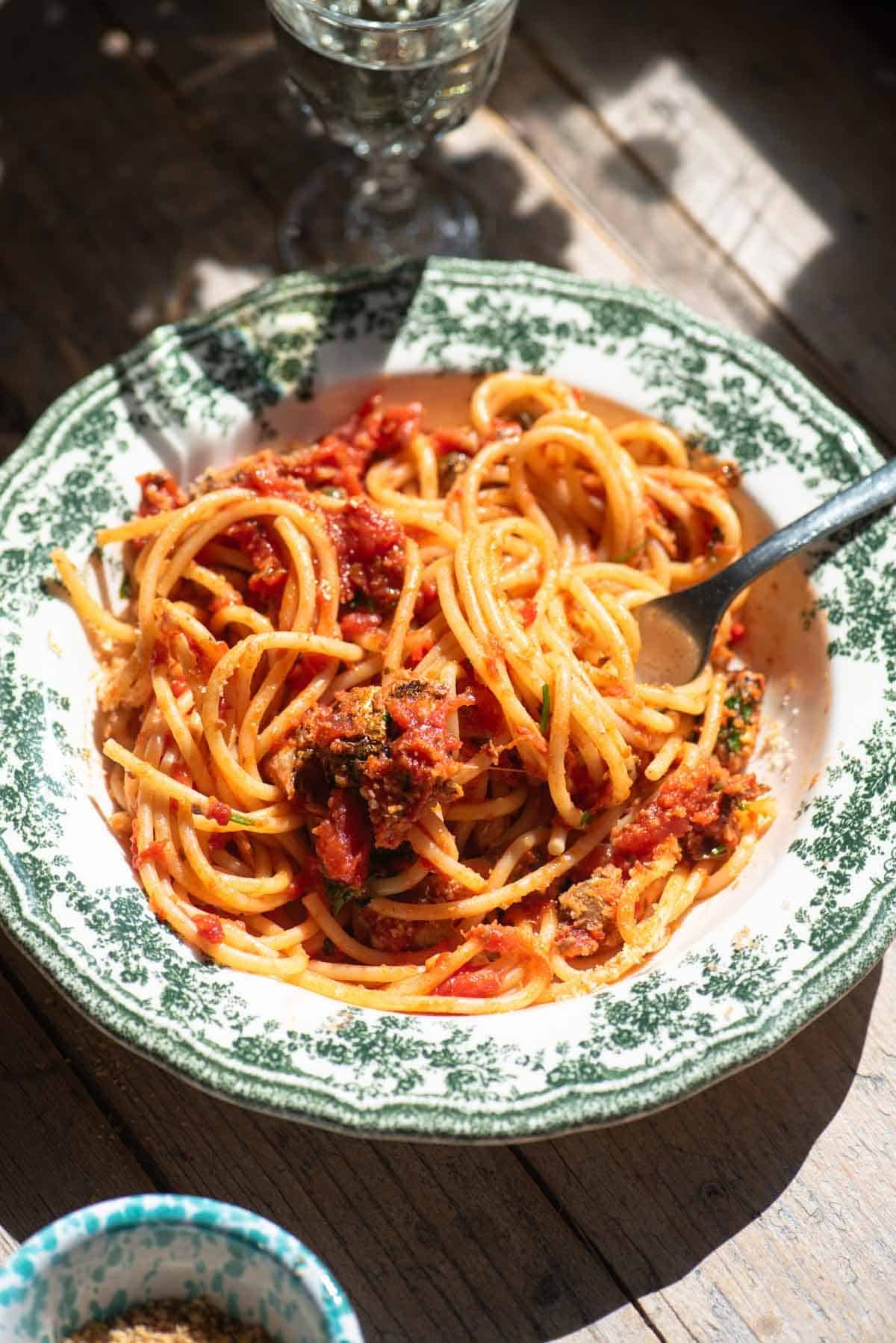 A side shot of tomato sardine spaghetti in a green and white bowl on a rustic wooden surface.