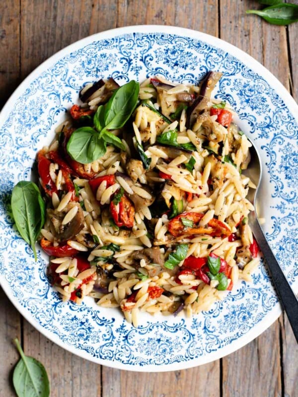 An orzo pasta salad with grilled veg in a blue bowl with a serving spoon at the side. The background is rustic wood.