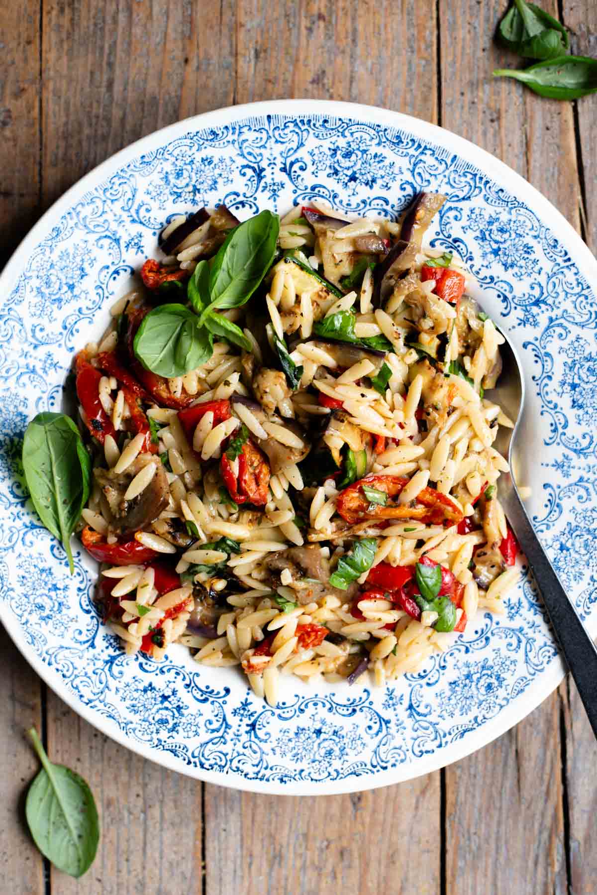 An orzo pasta salad with grilled veg in a blue bowl with a serving spoon at the side. The background is rustic wood.