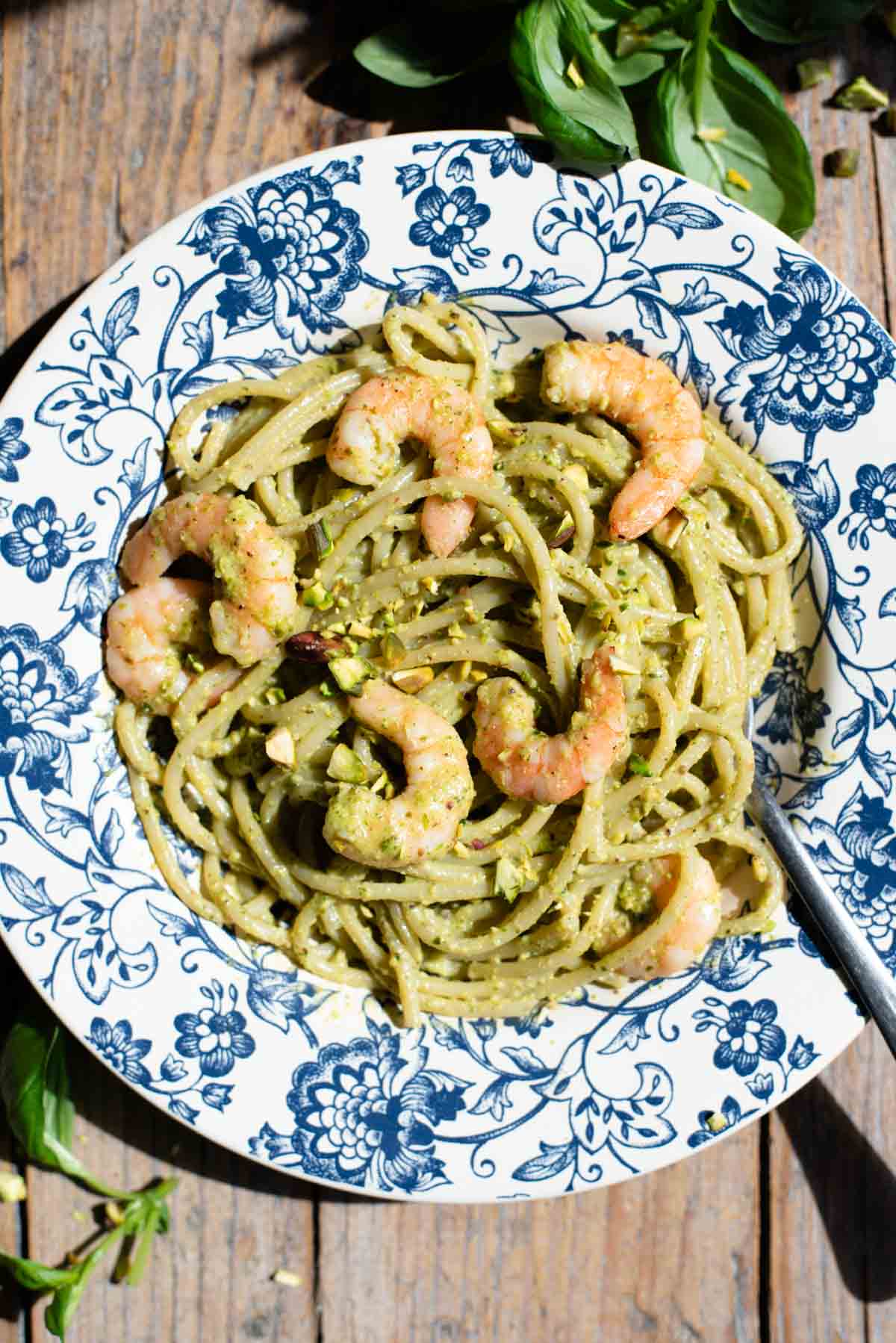 An overhead shot of pistachio pasta with shrimp in a blue floral bowl on a wooden background.