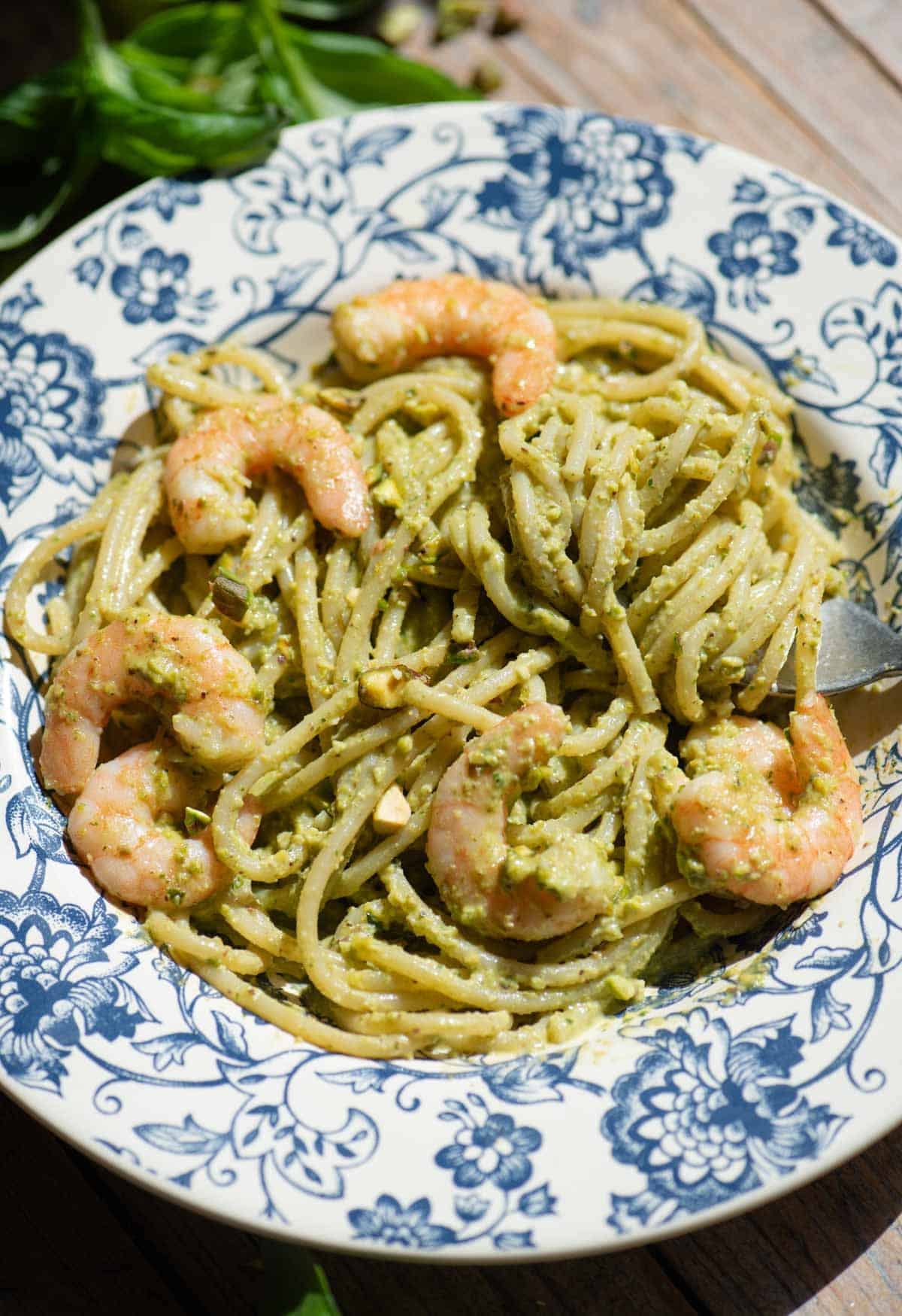A side shot of pistachio shrimp spaghetti in a blue bowl sitting on a wooden background.