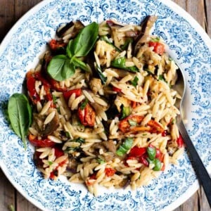 A close up of an orzo pasta salad with grilled veg and tomatoes in a blue bowl with basil on top.
