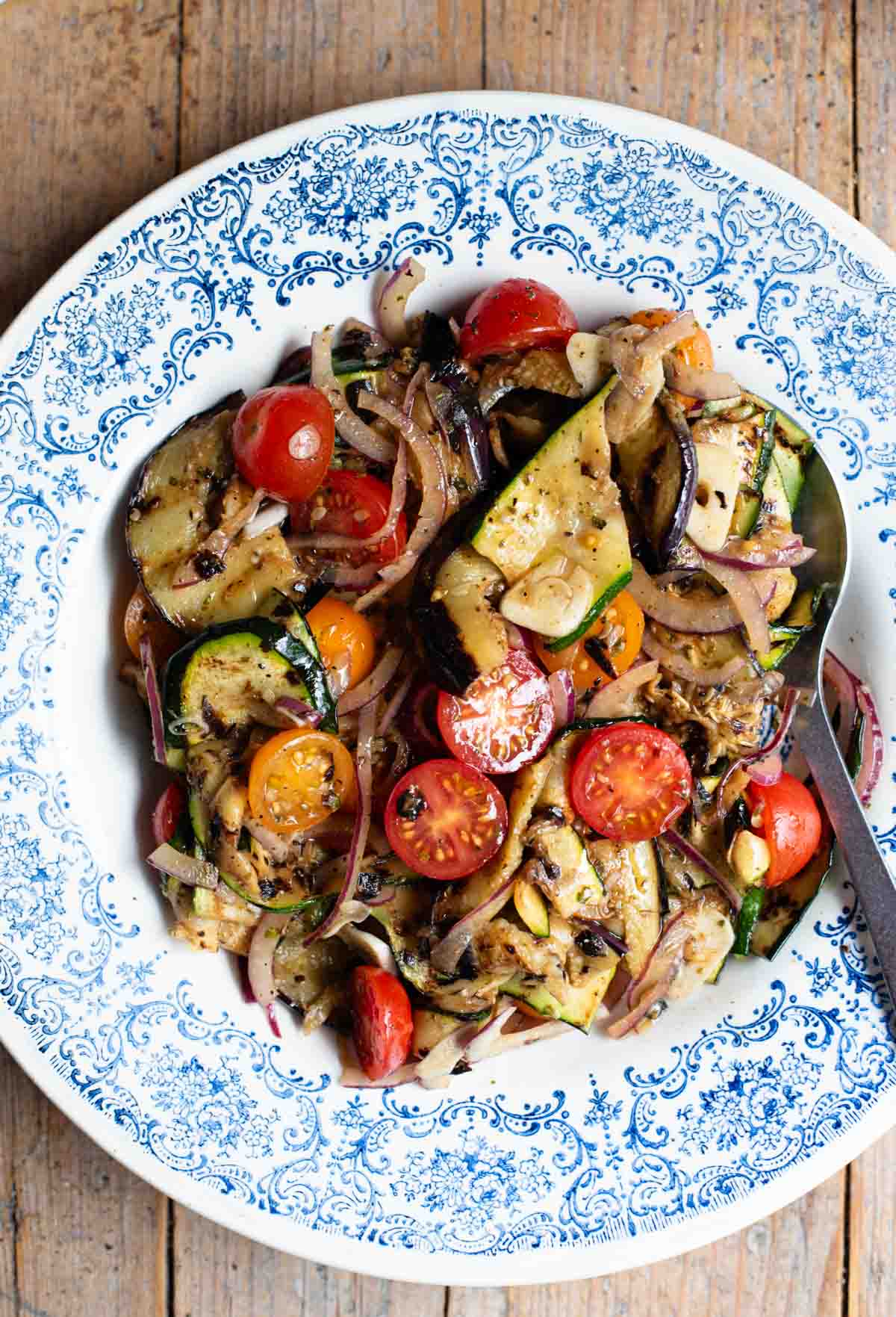 An overhead shot of marinated grilled summer vegetables in a blue bowl on a wooden surface.