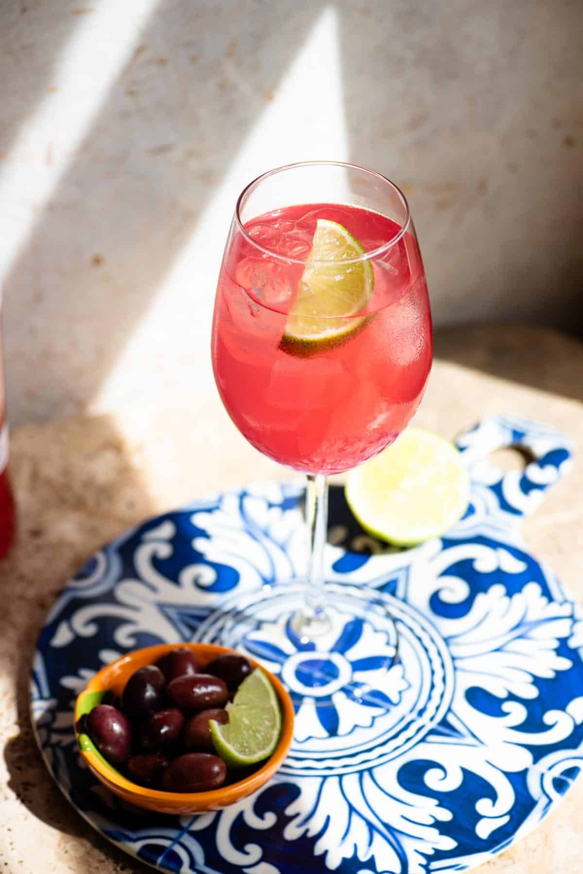 A close up of a sarti spritz with a wedge of lime on a blue patterned ceramic board with olives at the side.