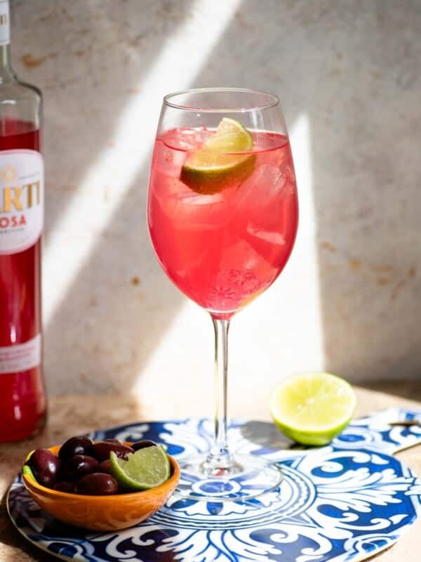A Sarti Spritz in a wine glass with ice and a slice of lime sitting on a blue ceramic serving board with olive at the side.