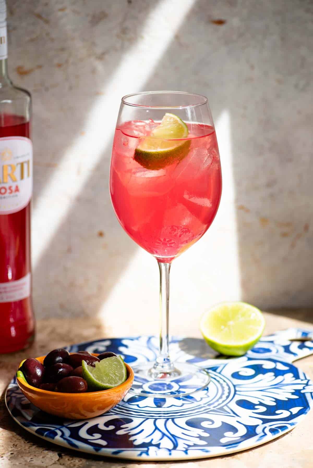 A Sarti Spritz in a wine glass with ice and a slice of lime sitting on a blue ceramic serving board with olive at the side.