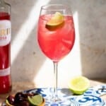 A Sarti Spritz in a tall wine glass garnished with a lime wedge. The cocktail is on a blue ceramic serving board with a small bowl of olives at the side.