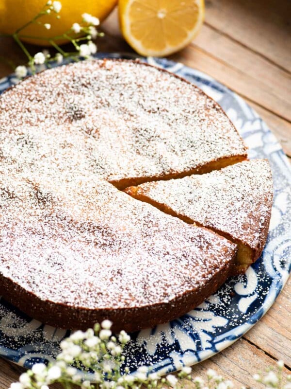 A white torta caprese on a blue ceramic board with fresh lemons behind it and small white flowers around.