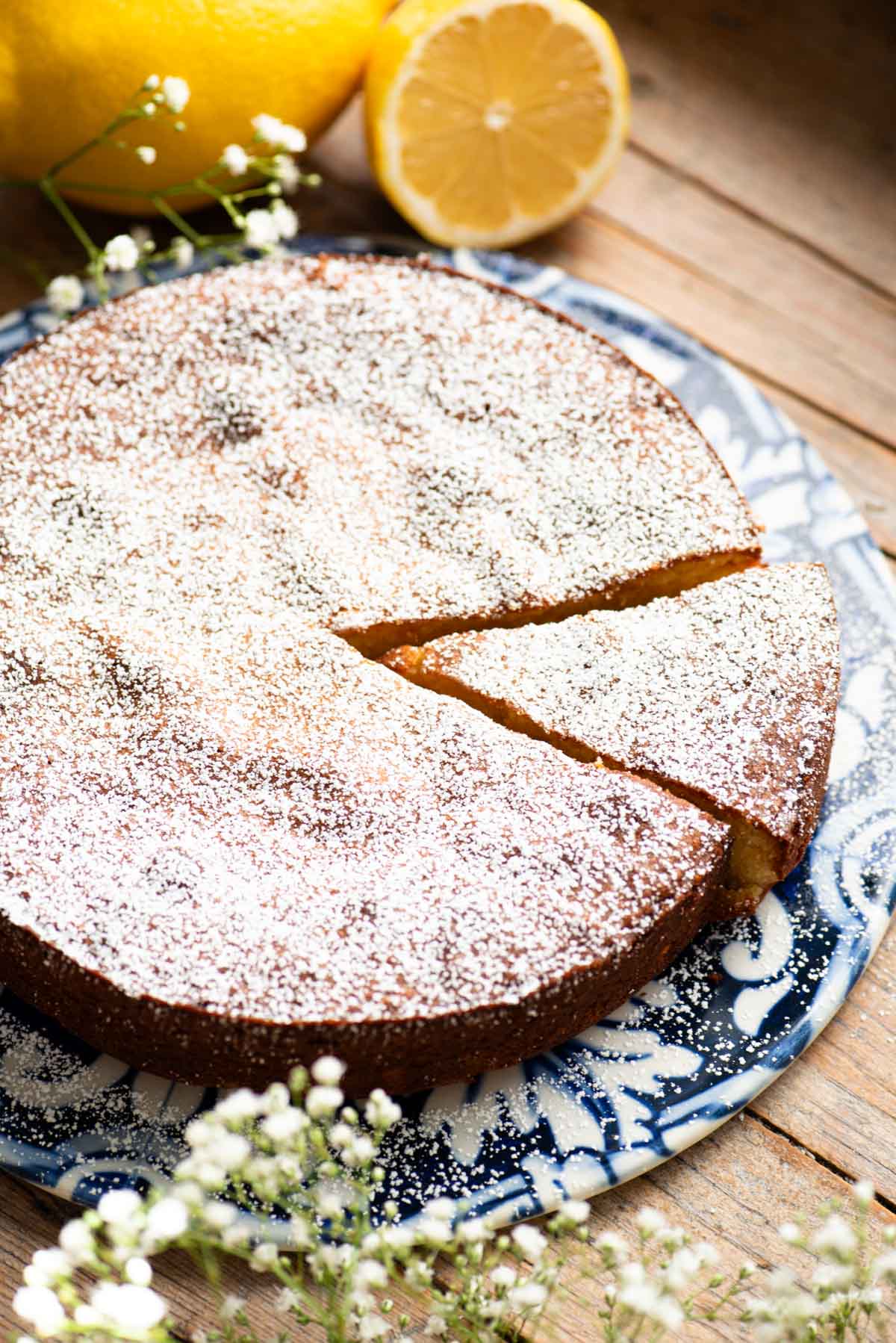 A white torta caprese on a blue ceramic board with fresh lemons behind it and small white flowers around.