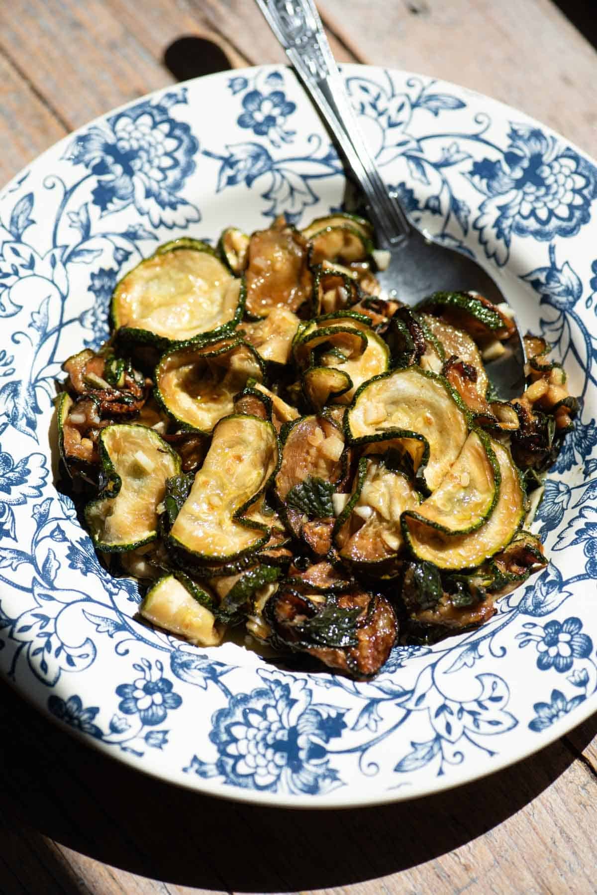 A blue and white floral bowl filled with fried marinated zucchini and a spoon.