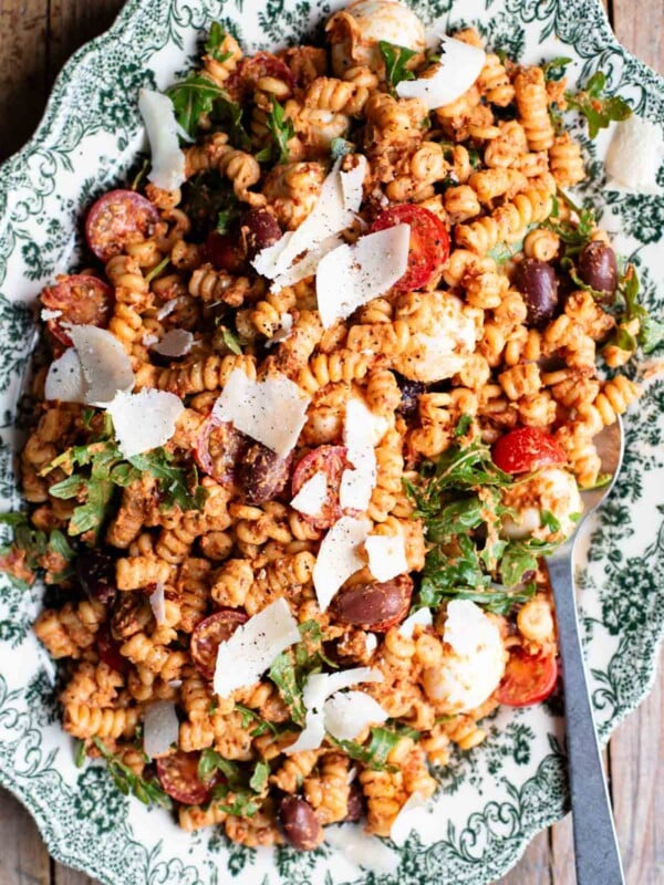 An overhead shot of a sundried tomato pasta salad on a serving plate with mozzarella, olives, arugula and parmigiano shavings.