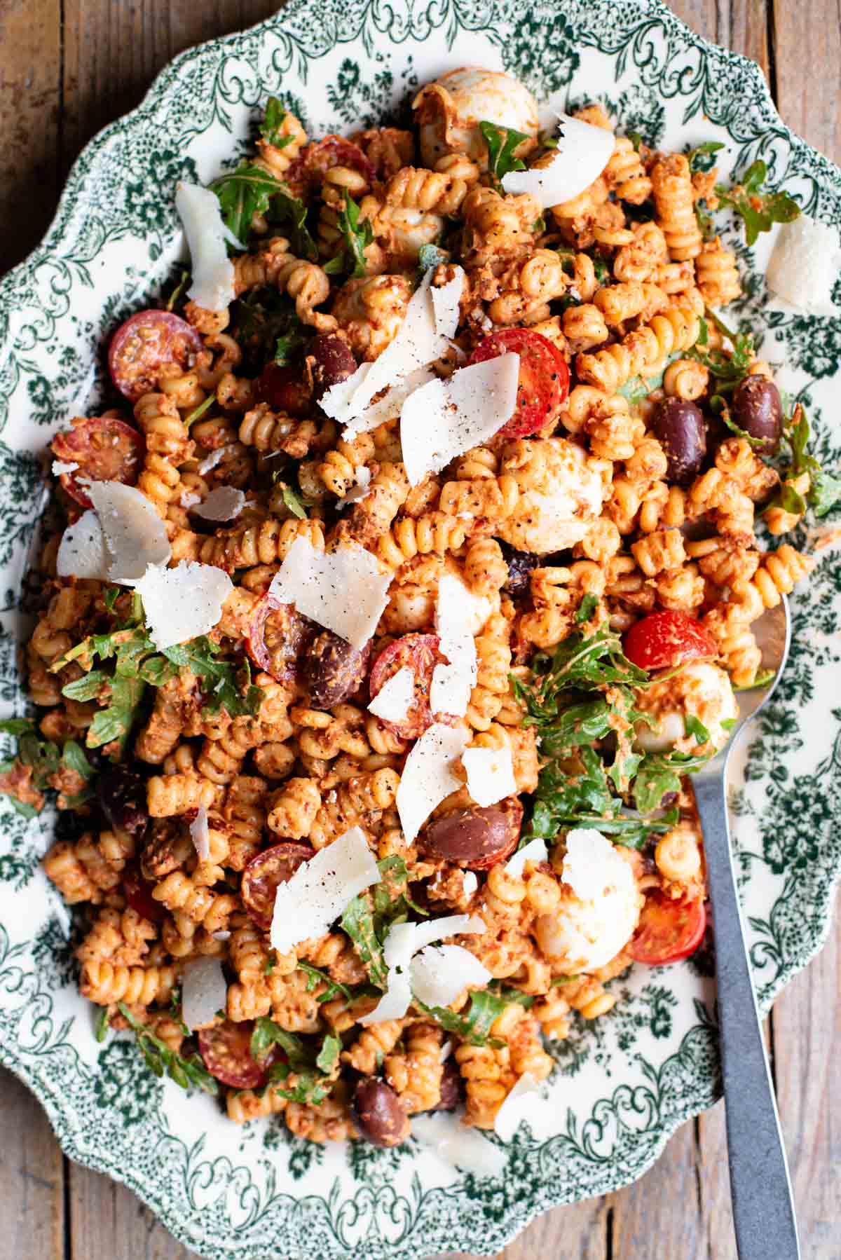 An overhead shot of a sundried tomato pasta salad on a serving plate with mozzarella, olives, arugula and parmigiano shavings.