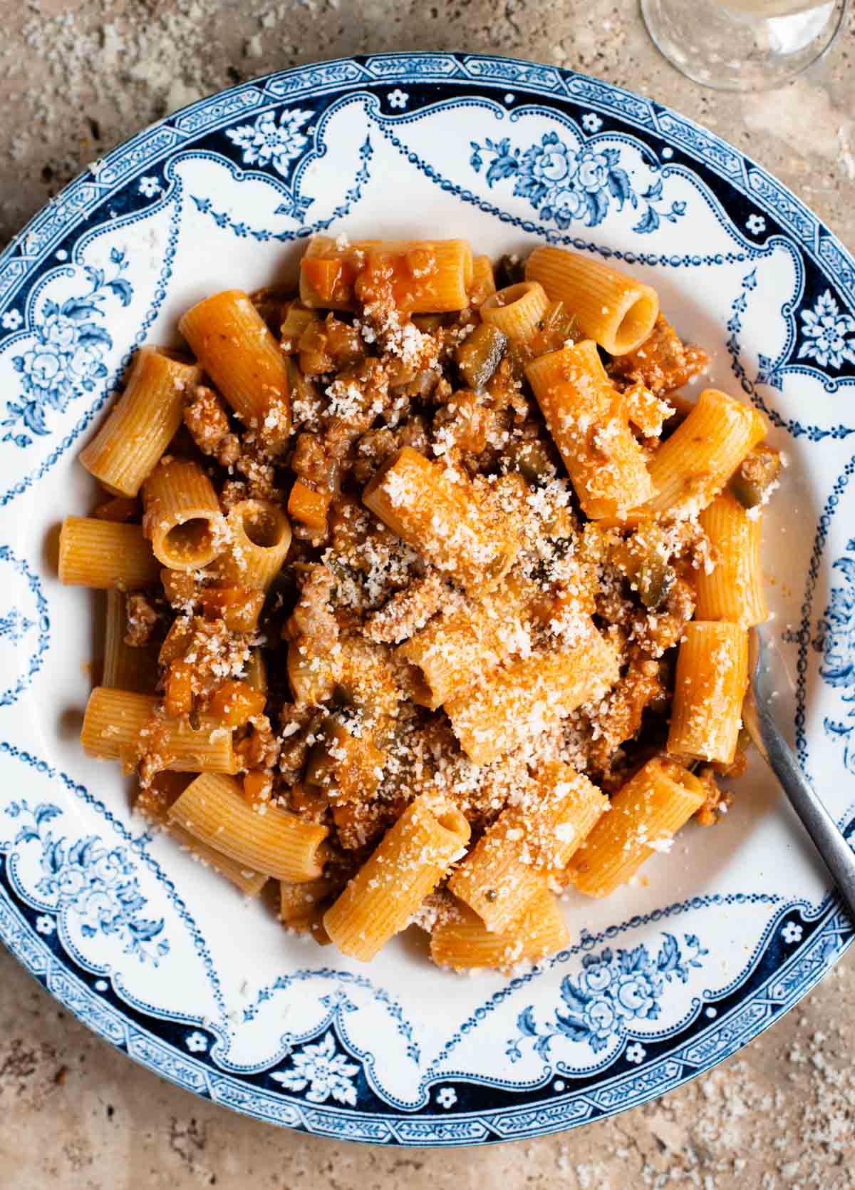 An overhead shot of rigatoni pasta with a sausage and eggplant ragu in a blue and white bowl on a travertine surface.