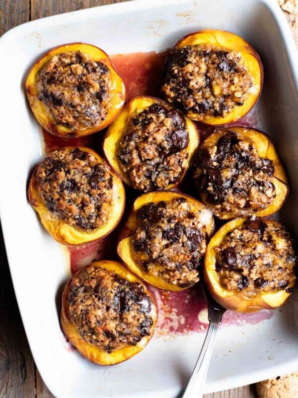 An overhead shot of baked peaches stuffed with amaretti cookies and chocolate in a white baking dish on a rustic wooden background.