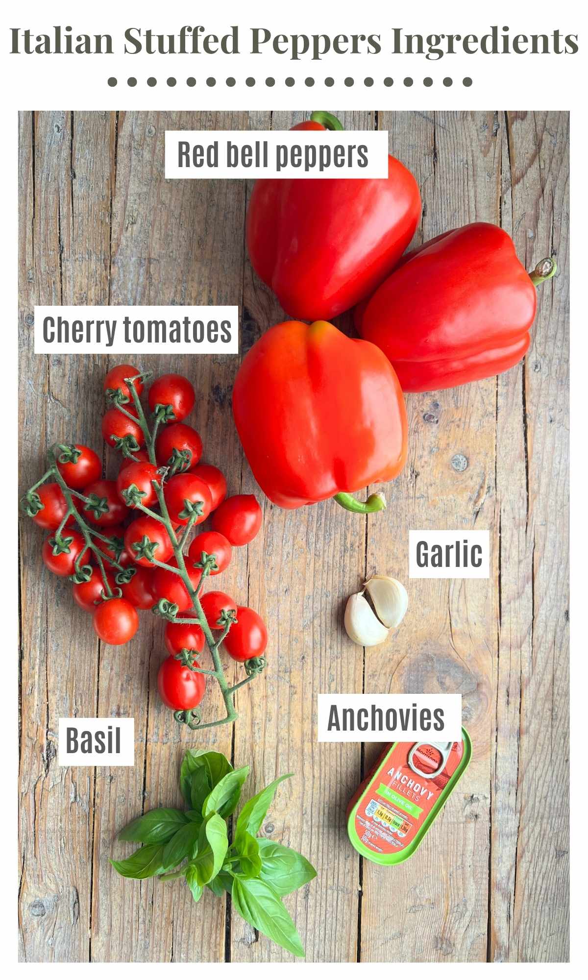An overhead shot of three red bell peppers, cherry tomatoes, garlic, basil and anchovies laying on rustic wooden background.