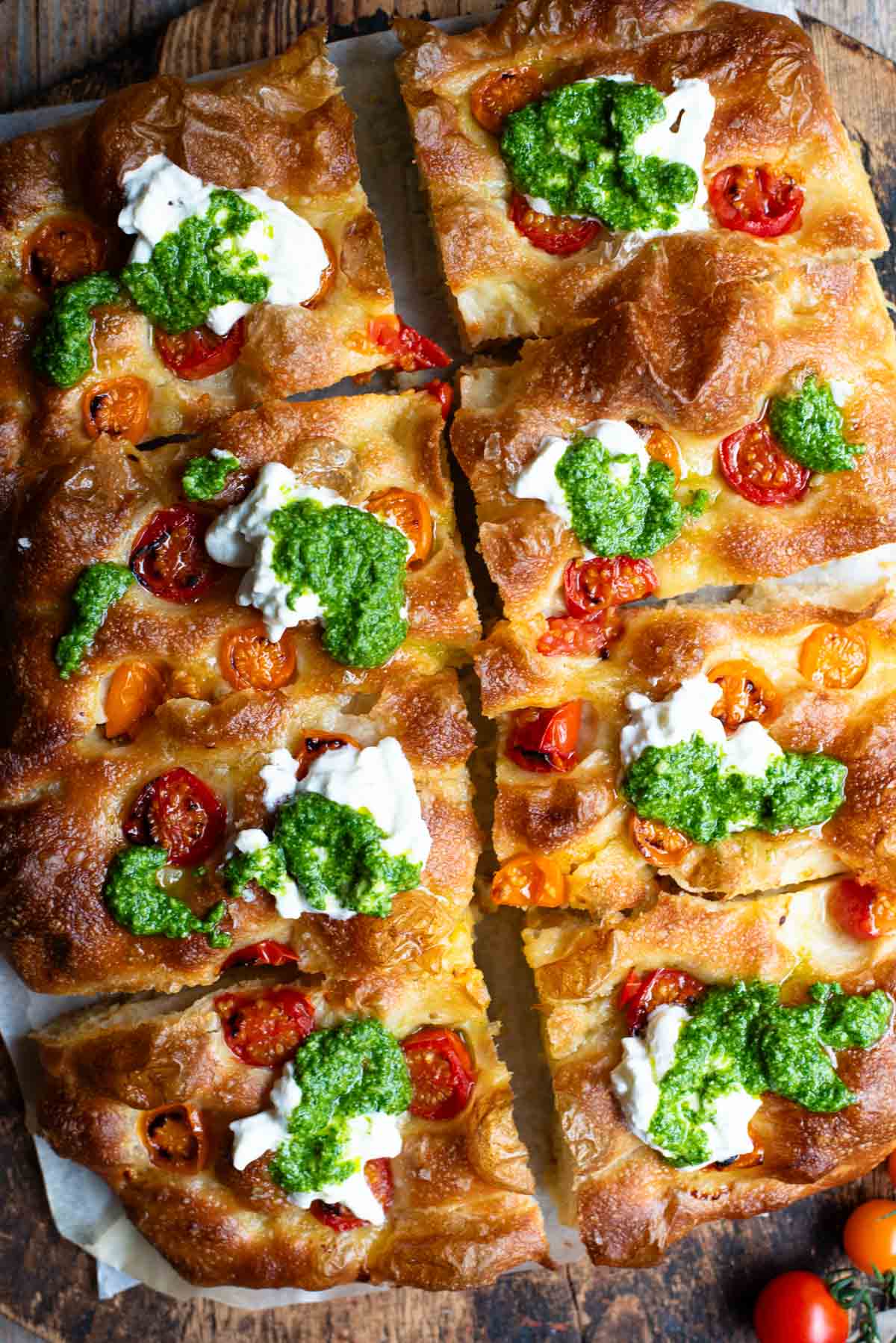 An overhead shot of a focaccia cut into 8 slices and topped with cherry tomatoes, pesto and burrata.