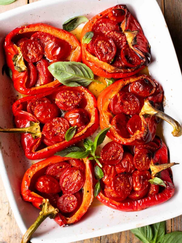 An overhead shot of 6 peppers in a white baking dish stuffed with cherry tomatoes, anchovies and garlic. The background is rustic wood.