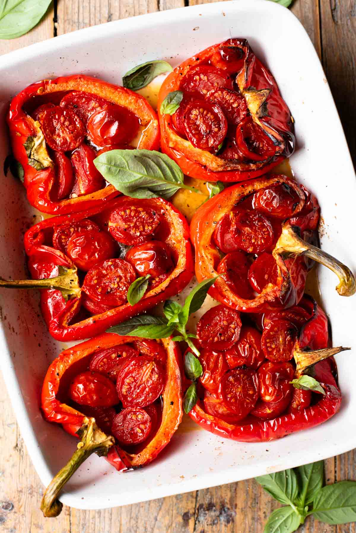 An overhead shot of 6 peppers in a white baking dish stuffed with cherry tomatoes, anchovies and garlic. The background is rustic wood.