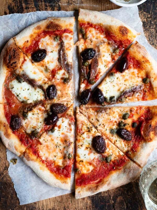An overhead shot of a pizza cut into slices on a rustic wooden board. The pizza is topped with olives, anchovies and capers.
