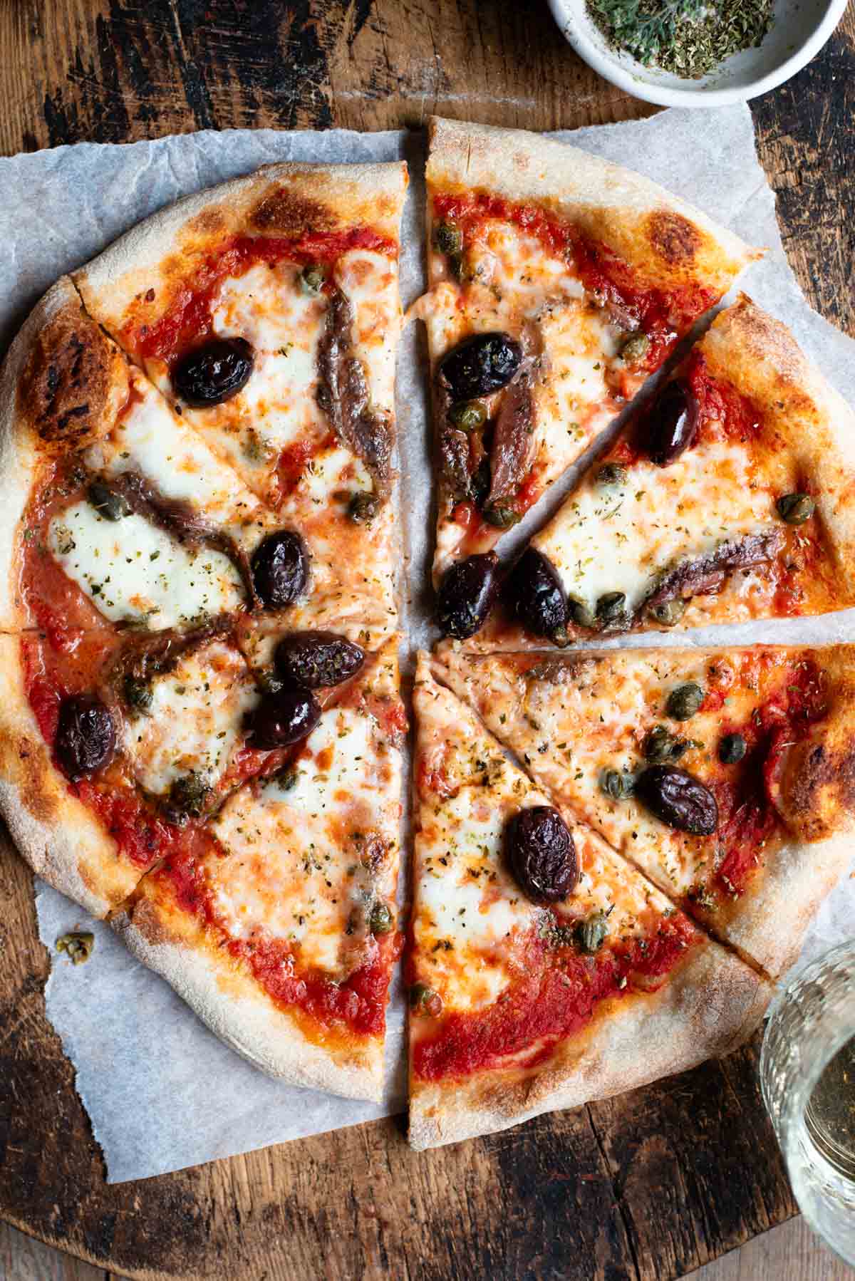 An overhead shot of a pizza cut into slices on a rustic wooden board. The pizza is topped with olives, anchovies and capers.