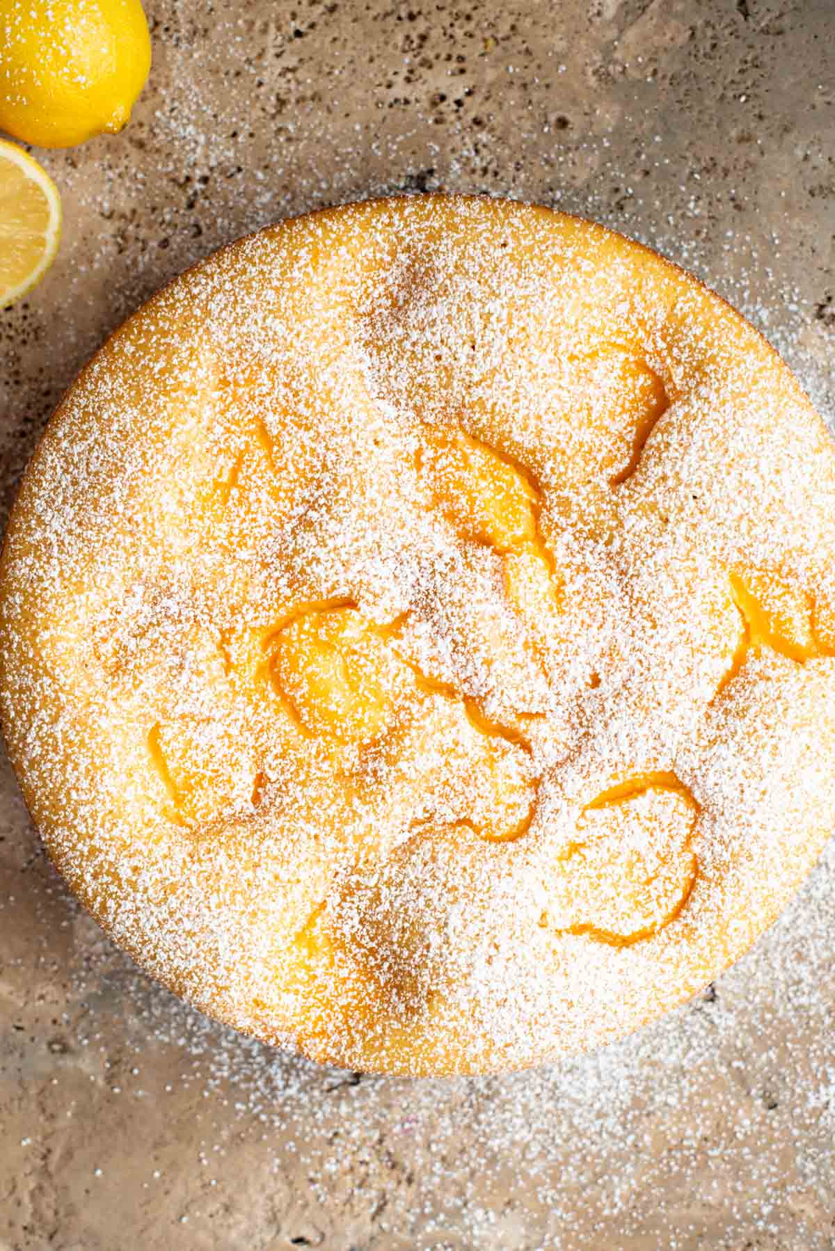 An overhead shot of a rustic Italian cake on a travertine surface with lemons at the side. The cake has dollops of pastry cream and powdered sugar.