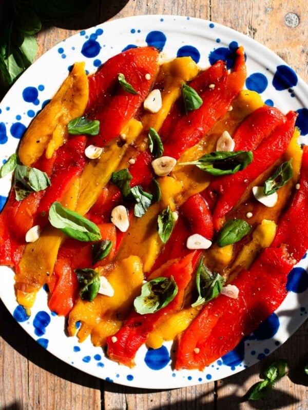 Red and yellow marinated roasted peppers arranged on a blue speckled plate topped with garlic and basil.
